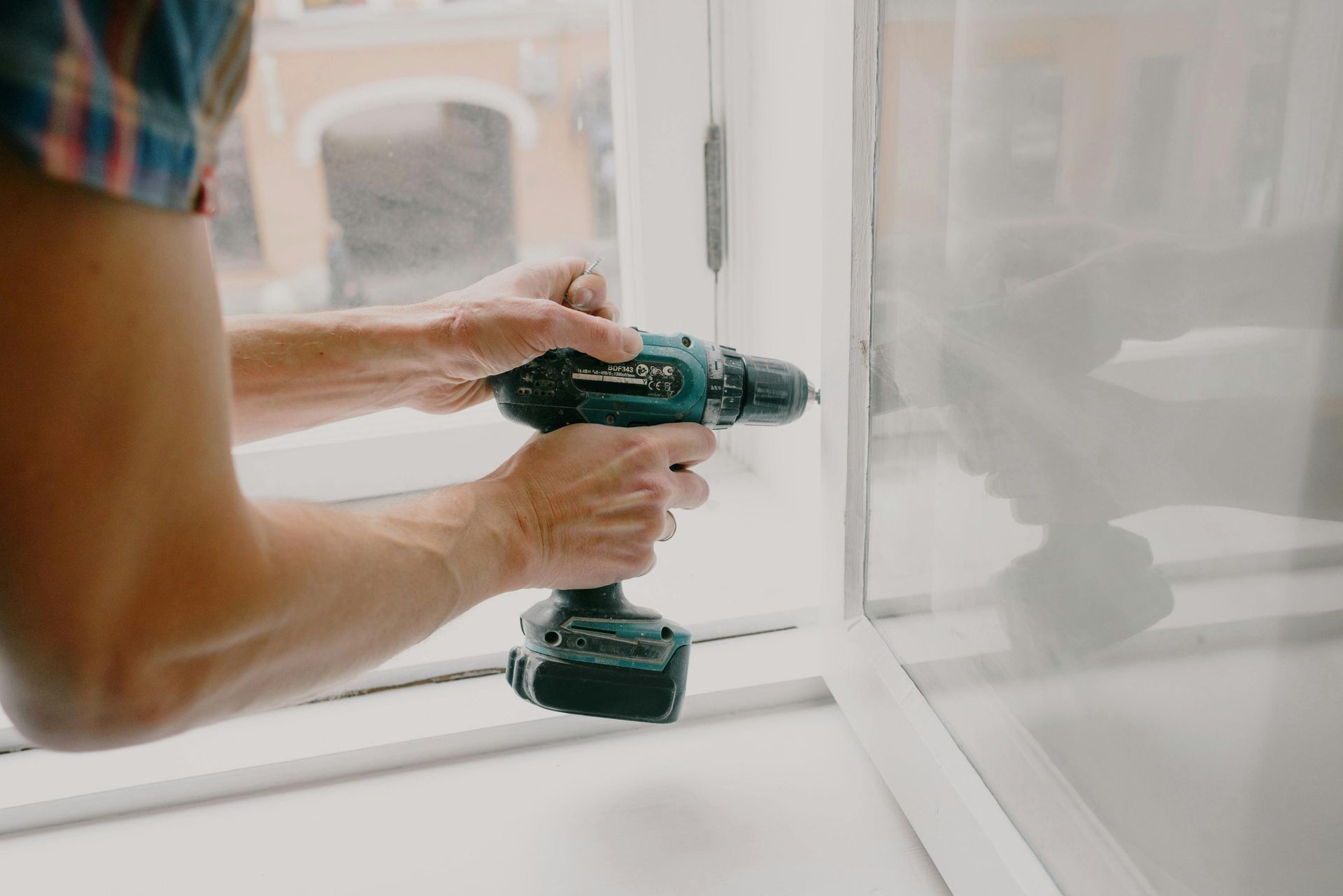 Person using a teal drill to work on a white window frame indoors.